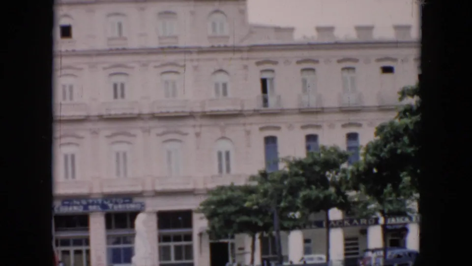1956 Cuba 8mm frame showing a pale classical building facade, leafy trees, and street-level signage partly obscured by dark foreground edges.