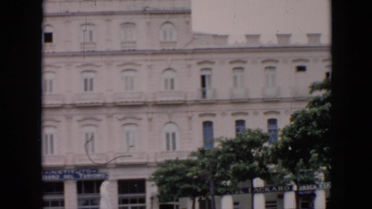 1956 Cuba frame of a large pale building with repeated windows, trees in front, and partially legible tourism or hotel signage at street level.