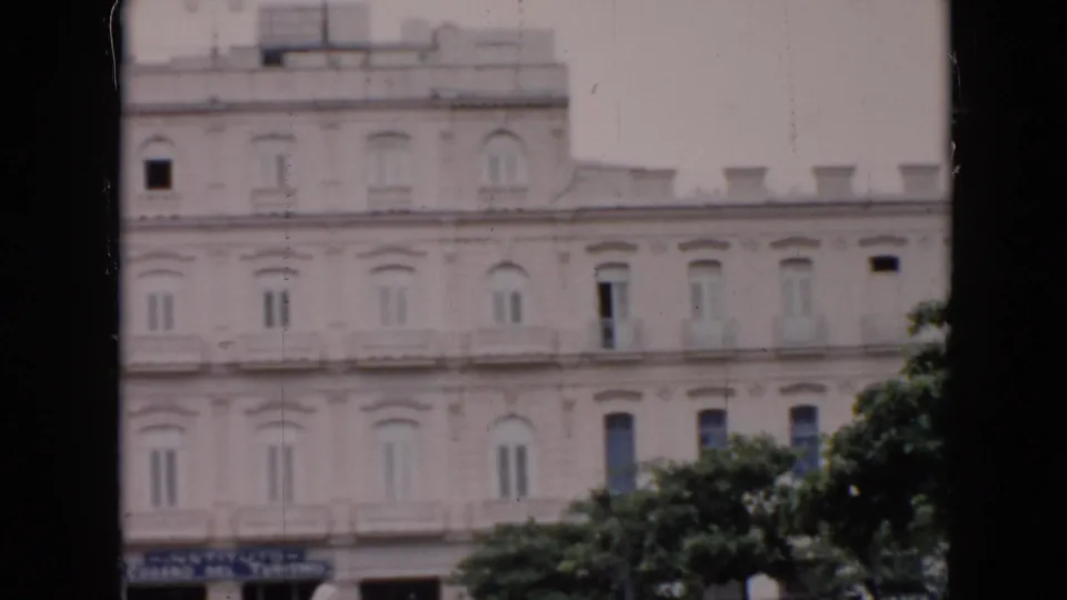 1956 Cuba 8mm frame showing the lower portion of a white multi-story building, tree canopy, and blue signs along the ground floor.