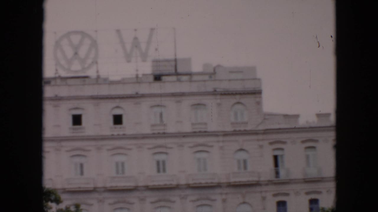 1956 Cuba 8mm frame showing a pale ornate building with arched windows and a rooftop Volkswagen sign, viewed through dark foreground bars.