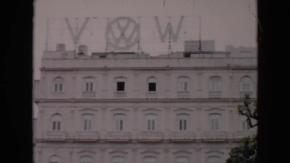 1956 Cuba 8mm still showing an ornate pale building and large rooftop Volkswagen sign, with dark foreground framing and slight color fading.