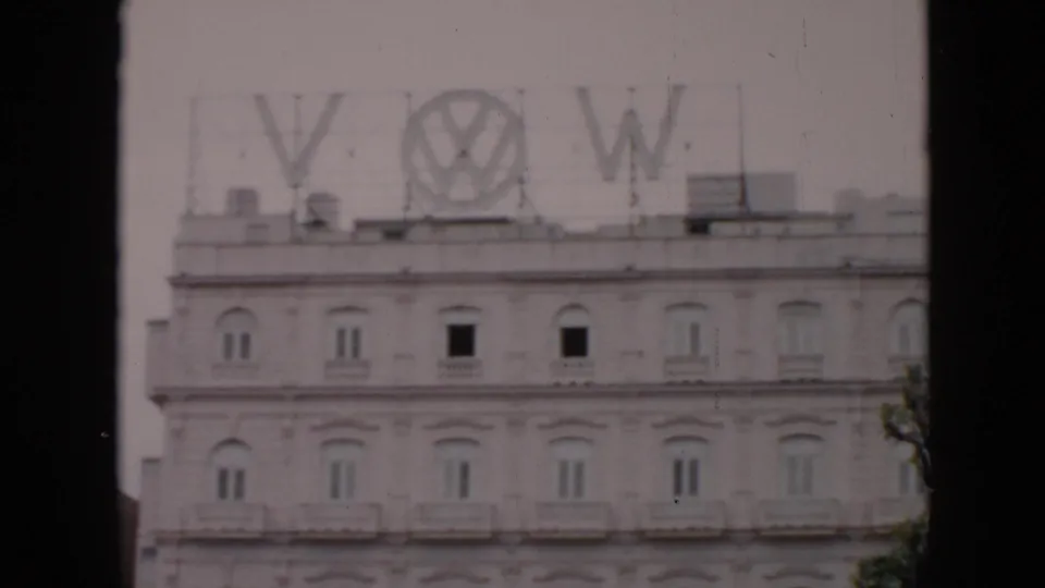 Final 1956 Cuba frame of the pale multi-story building with rooftop VW branding, arched windows, and dark vertical framing at the edges.