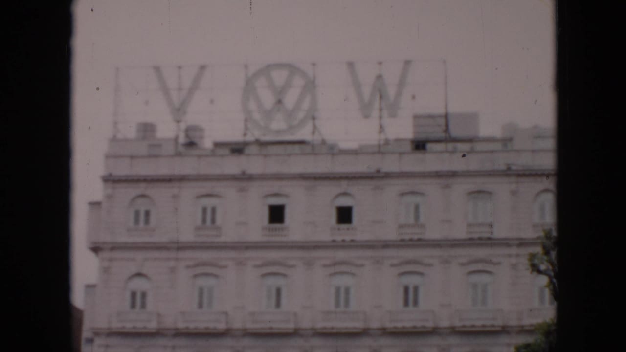 1956 Cuba 8mm frame showing a pale multi-story building with large rooftop VW and Volkswagen letters, filmed from across the street.