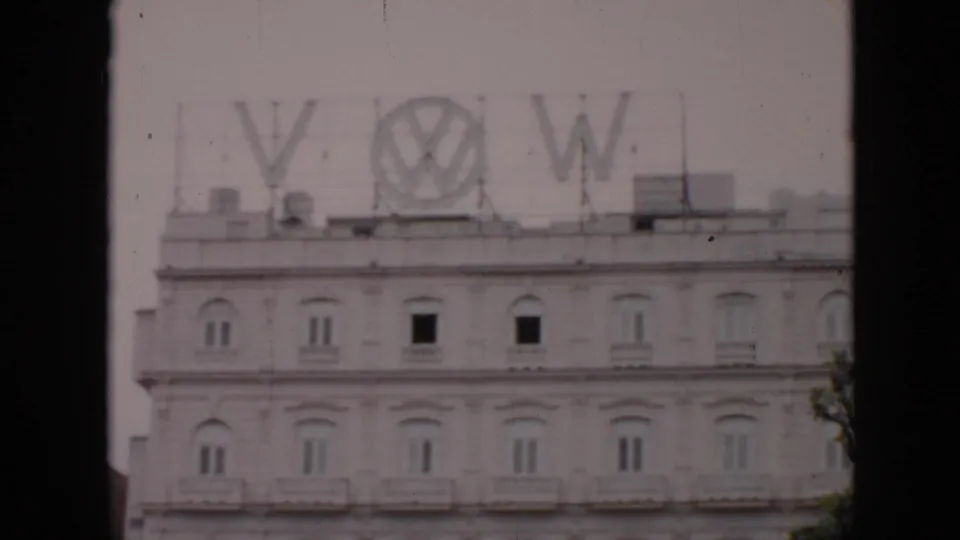 1956 Cuba 8mm frame showing a pale multi-story building with large rooftop VW and Volkswagen letters, filmed from across the street.