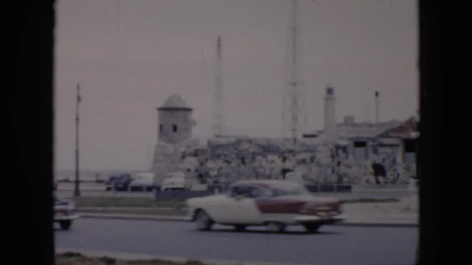 A 1956 Cuba street view shows mid-century cars moving past stone walls, a small tower, poles, and low waterfront structures.