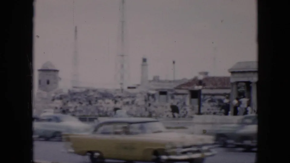 1956 Cuba 8mm frame with several moving mid-century cars, stone fortifications, a small tower, and people near a roadside shelter.