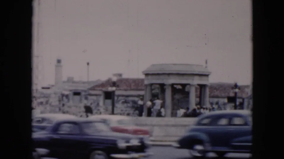 A 1956 Cuba roadway scene shows mid-century cars passing pedestrians beside a shelter, stone masonry, and a small tower or lighthouse.