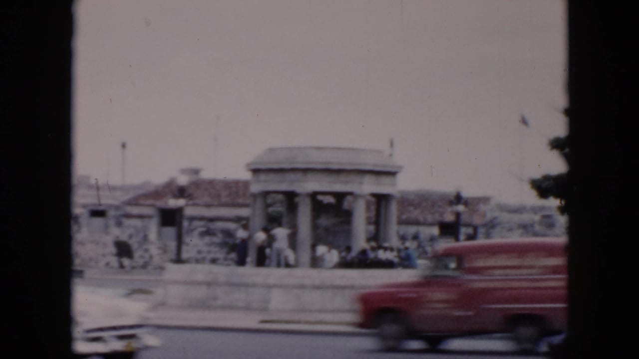 1956 Cuba street view with a red vehicle and other cars passing a stone columned shelter where pedestrians stand nearby.