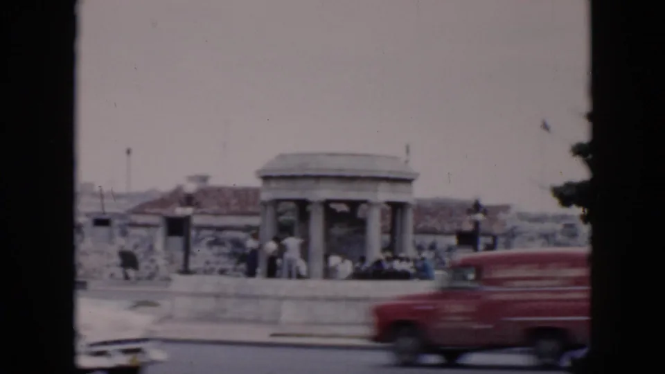 1956 Cuba street view with a red vehicle and other cars passing a stone columned shelter where pedestrians stand nearby.