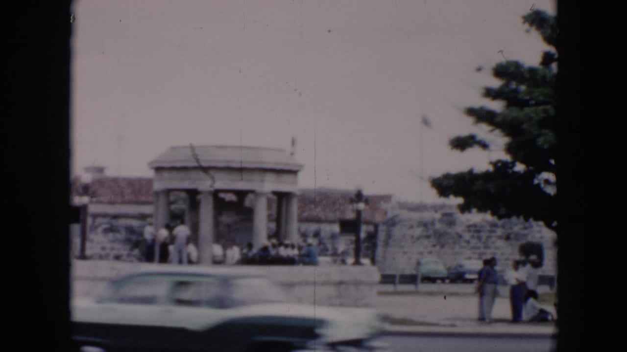 A light-colored mid-century car moves across a Cuba street scene with pedestrians near a stone shelter and low wall.