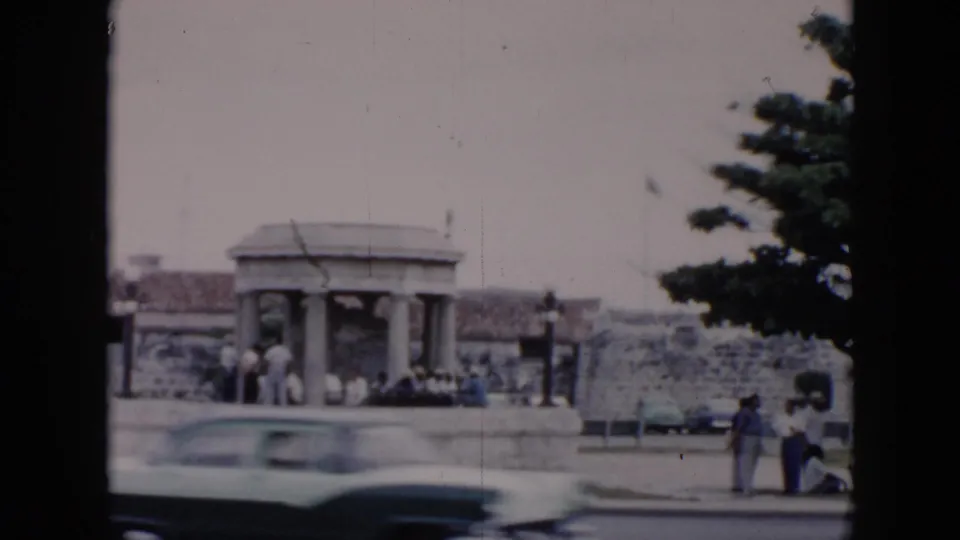A light-colored mid-century car moves across a Cuba street scene with pedestrians near a stone shelter and low wall.