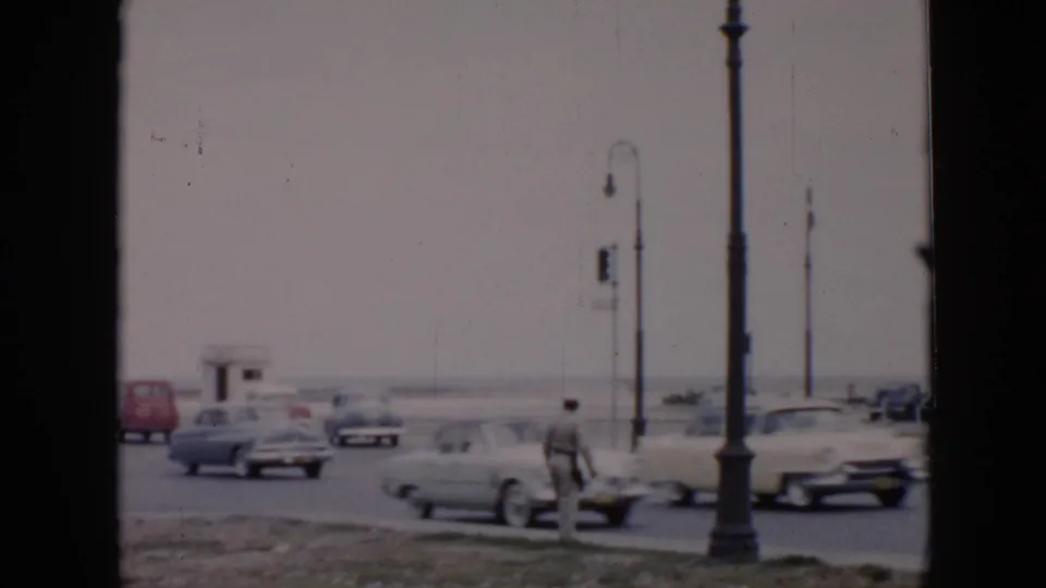 A broad 1956 Cuba roadway shows several mid-century cars, tall street lamps, and a pedestrian standing near the curb.