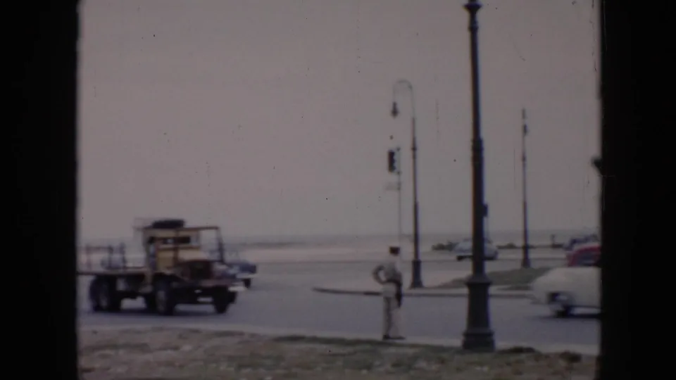 A utility truck and cars pass along a Cuba road with tall lamps, a pedestrian at the roadside, and an open distant horizon.