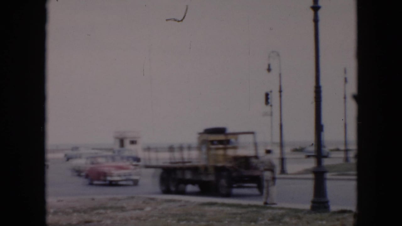 City intersection with classic cars, a yellow flatbed truck, street lamps, and a traffic officer.