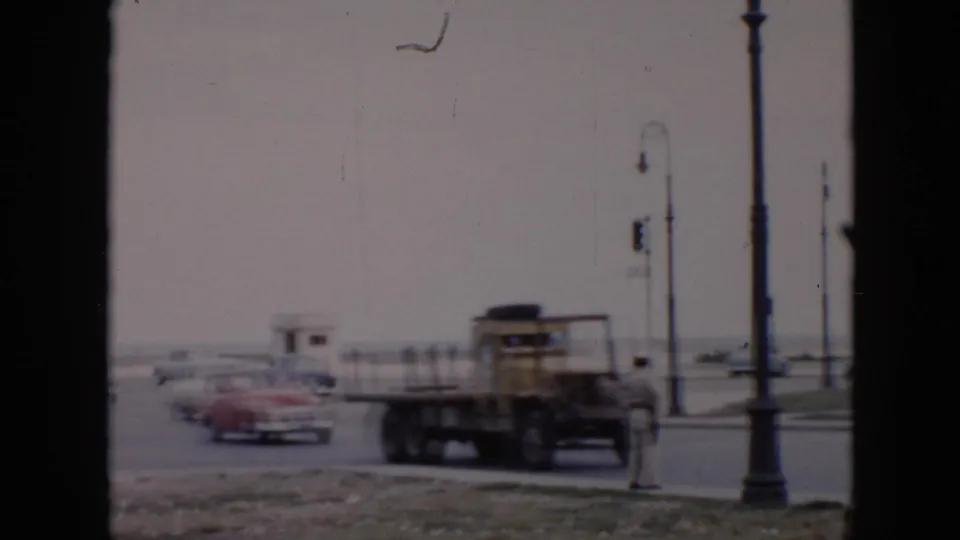 Mid-1950s Cuba street view with classic cars, a yellow flatbed truck, tall lamps, and a uniformed traffic officer near an intersection.
