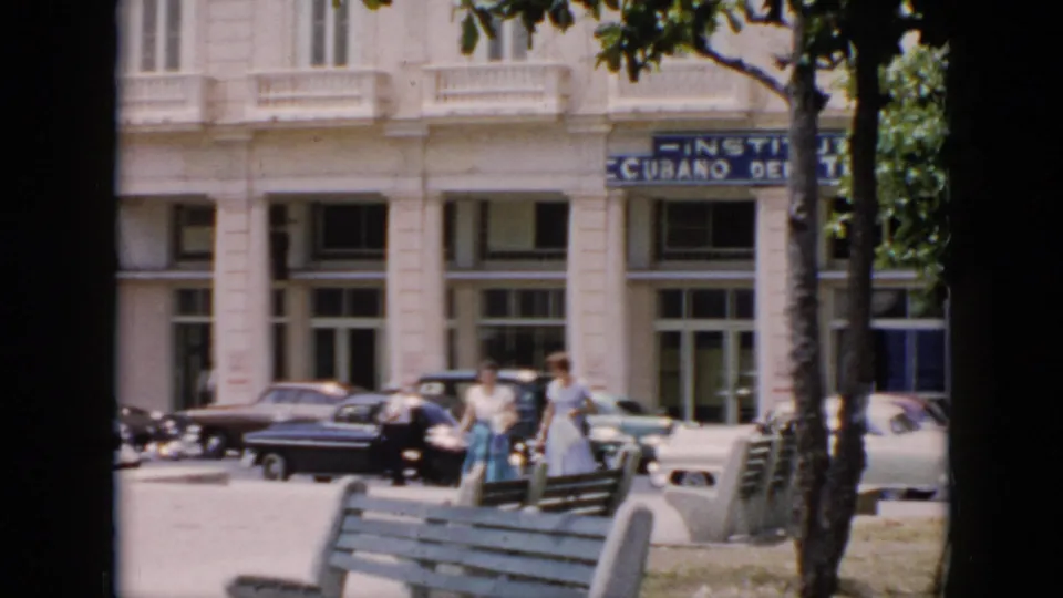 Mid-1950s Cuba street-side view with pedestrians walking past benches, trees, parked classic cars, and a large facade with partial signage.