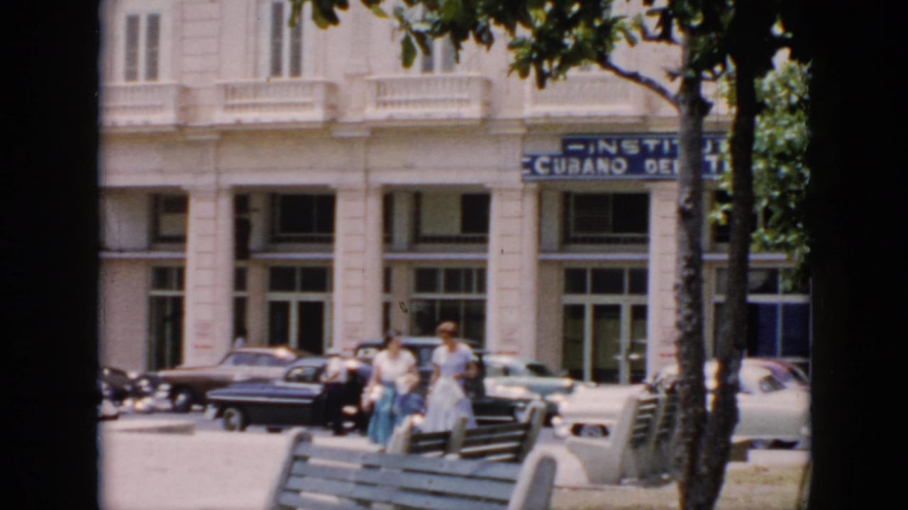 1956 Cuba street view with two women walking past benches and classic cars in front of a white building with partial Instituto Cubano signage.