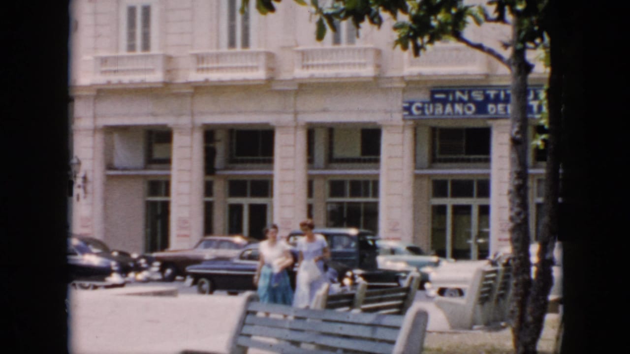 Two women in 1950s clothing walk through a sunlit Cuba street scene with benches, trees, parked cars, and a white institutional facade.