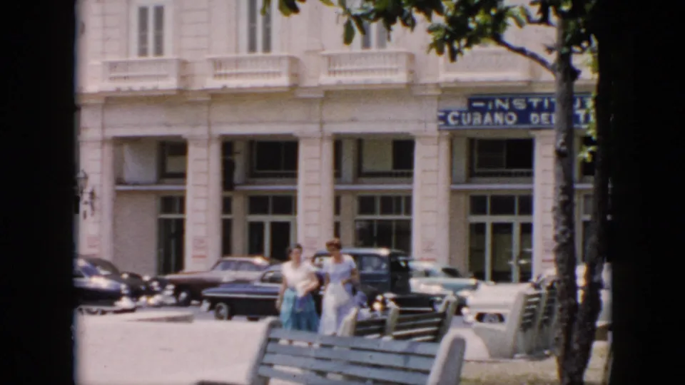 Two women in 1950s clothing walk through a sunlit Cuba street scene with benches, trees, parked cars, and a white institutional facade.