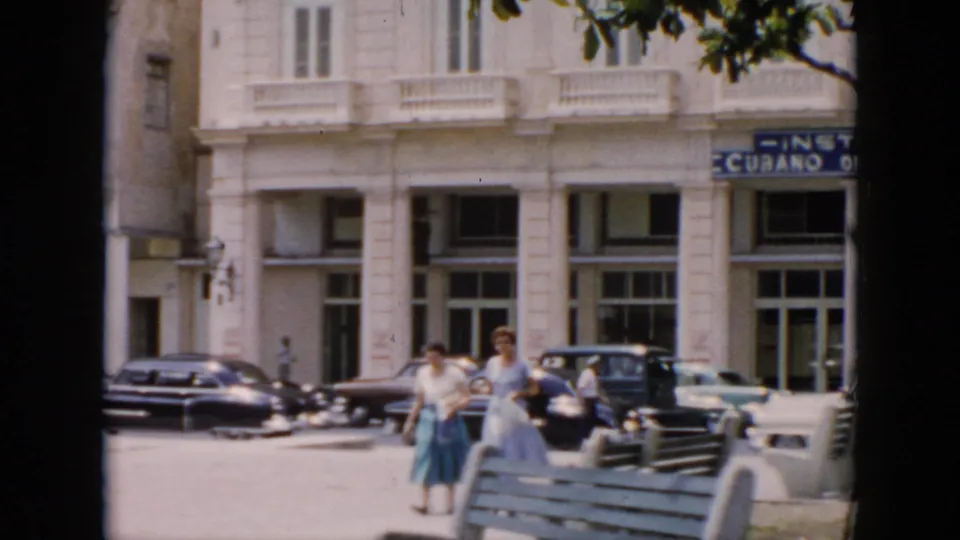 A 1956 Cuba sidewalk shot shows two women approaching the camera near a bench, with parked cars and a white multi-story facade behind them.