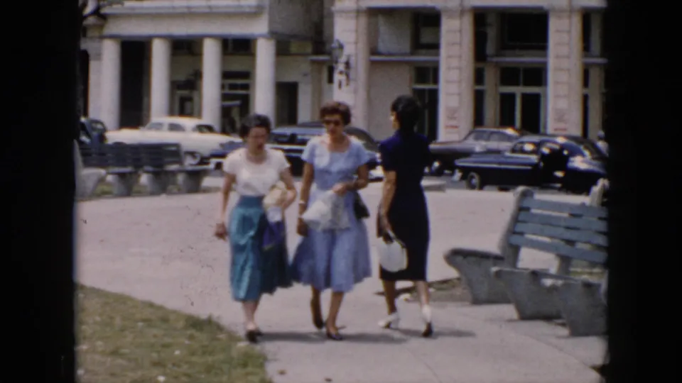Three women walk along a paved path in a 1956 Cuba street scene, with benches, grass, parked cars, and a white building entrance nearby.