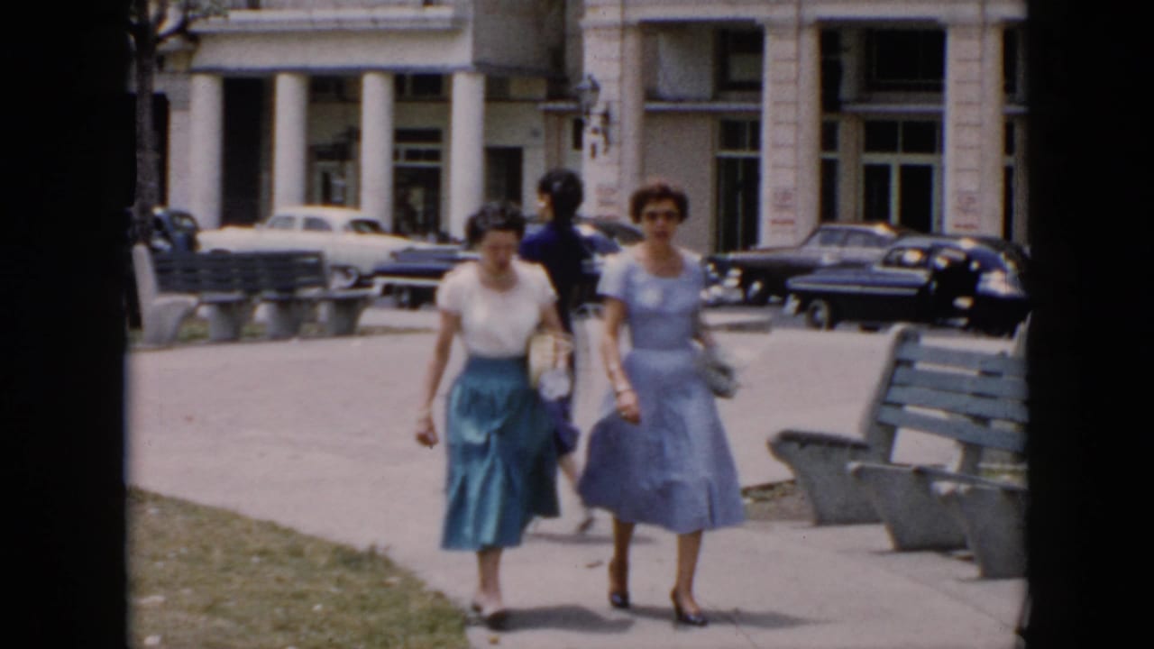 In 1956 Cuba, two women in mid-century clothing walk along a city sidewalk past benches, grass, classic cars, and a columned building.