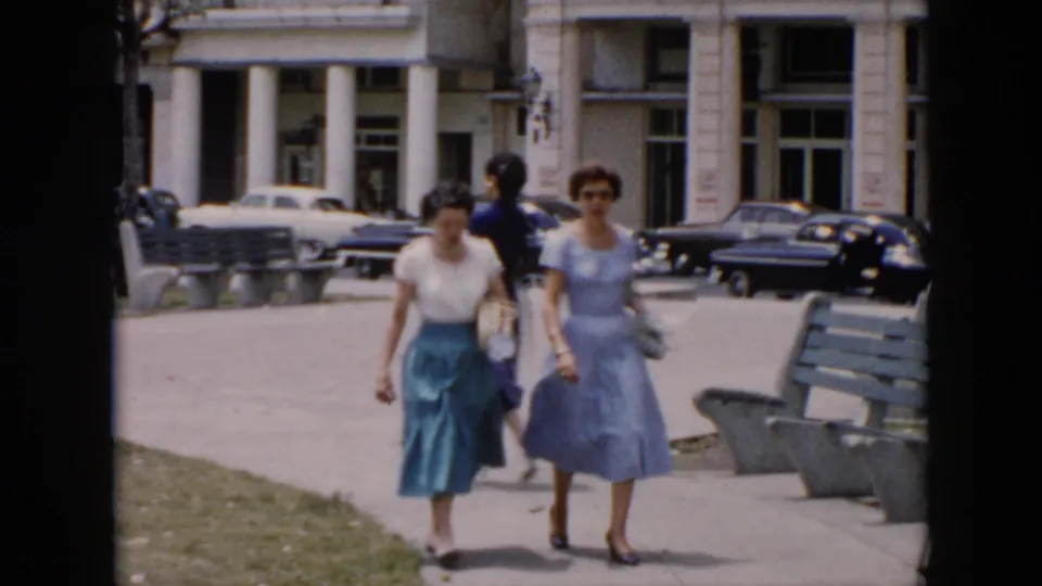 In 1956 Cuba, two women in mid-century clothing walk along a city sidewalk past benches, grass, classic cars, and a columned building.