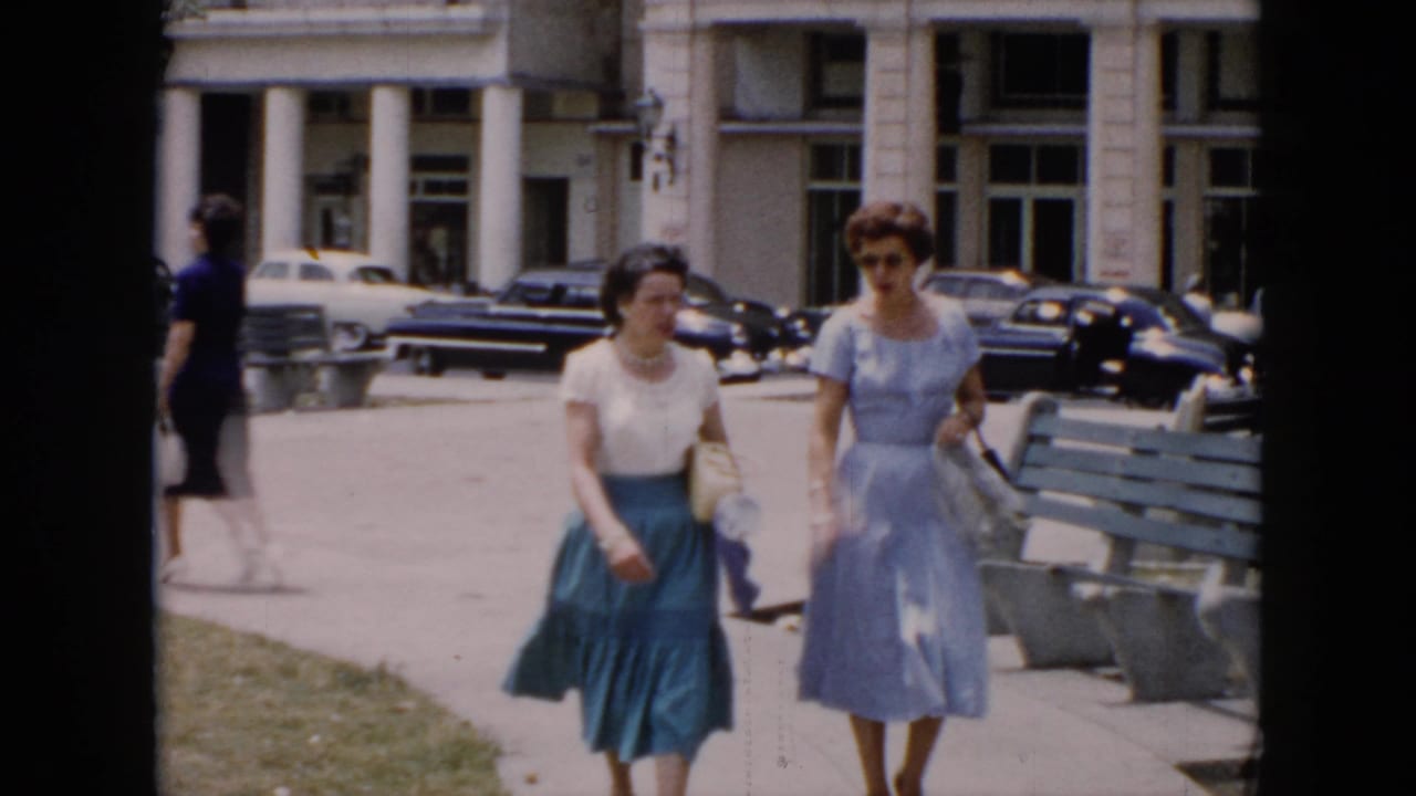 Two women continue walking past benches with classic cars and a building behind them.