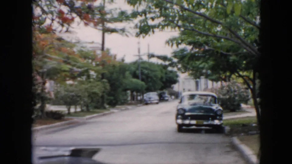 1956 Cuba 8mm frame showing a dark mid-century car near the curb, leafy branches overhead, lawns, and a quiet residential roadway.