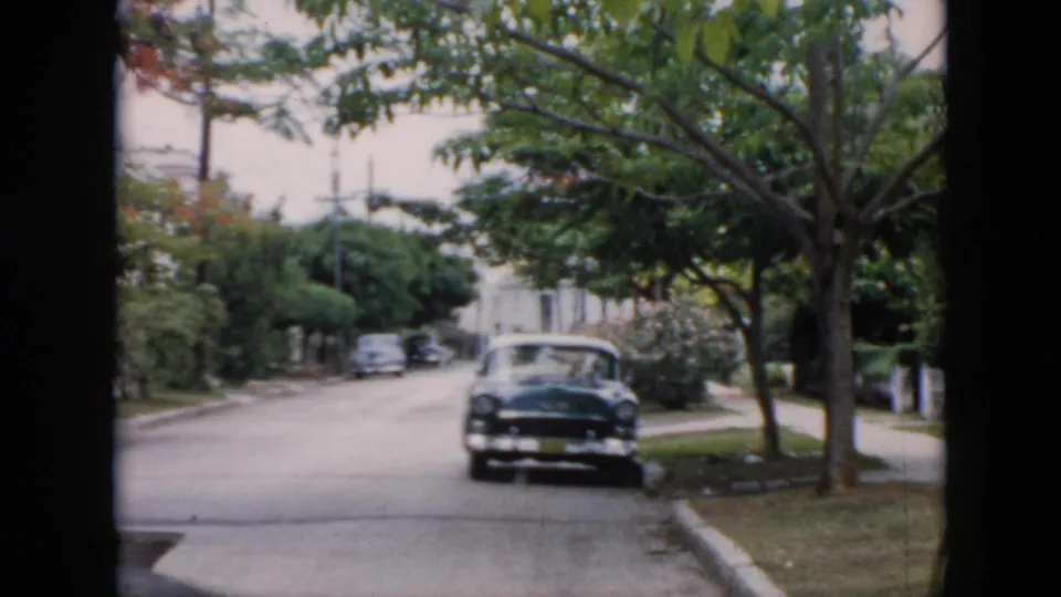 1956 Cuba residential street scene with a dark parked car, overhanging trees, sidewalks, lawns, and softly faded 8mm color.