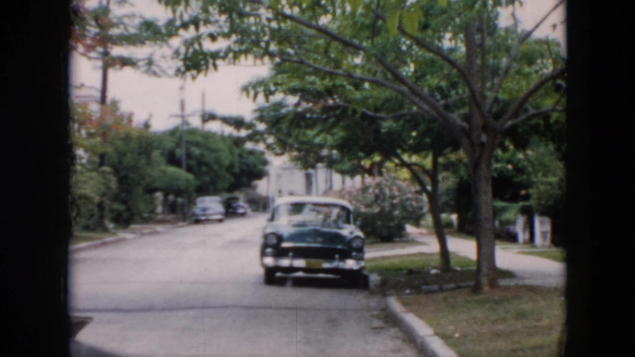 1956 Cuba home-movie frame showing a parked mid-century car on a tree-lined residential street with lawns and sidewalks.