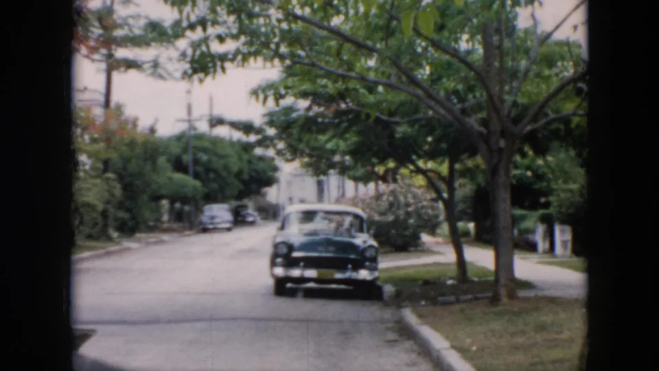 1956 Cuba home-movie frame showing a parked mid-century car on a tree-lined residential street with lawns and sidewalks.