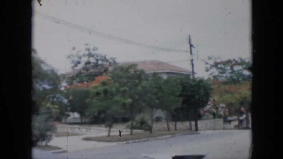 1956 Cuba home-movie frame of a residential avenue bordered by trees, low walls, tiled-roof houses, and overhead utility lines.
