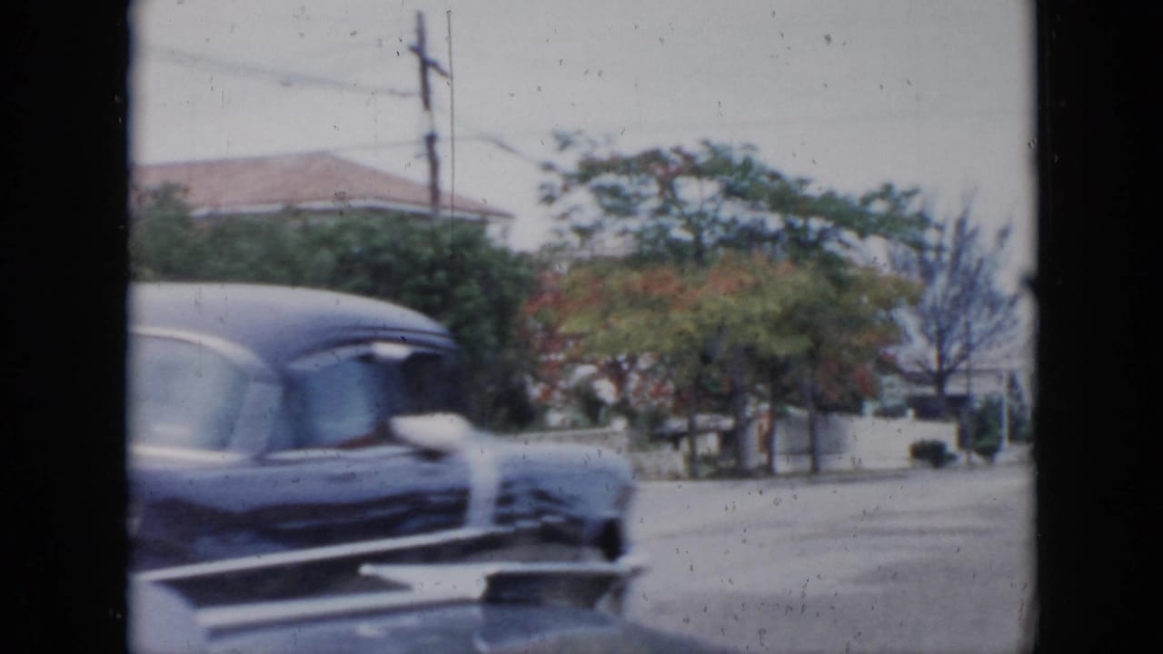 Dark blue mid-century car passing in front of trees and walled houses.