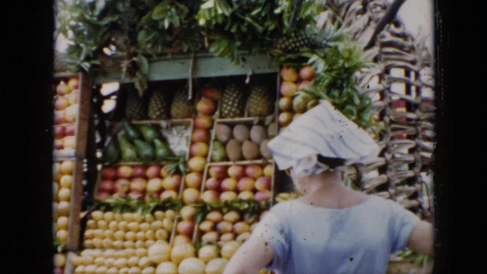 Open-air fruit stand in Cuba, 1956, with stacked produce and a shopper seen from behind wearing a white head covering.