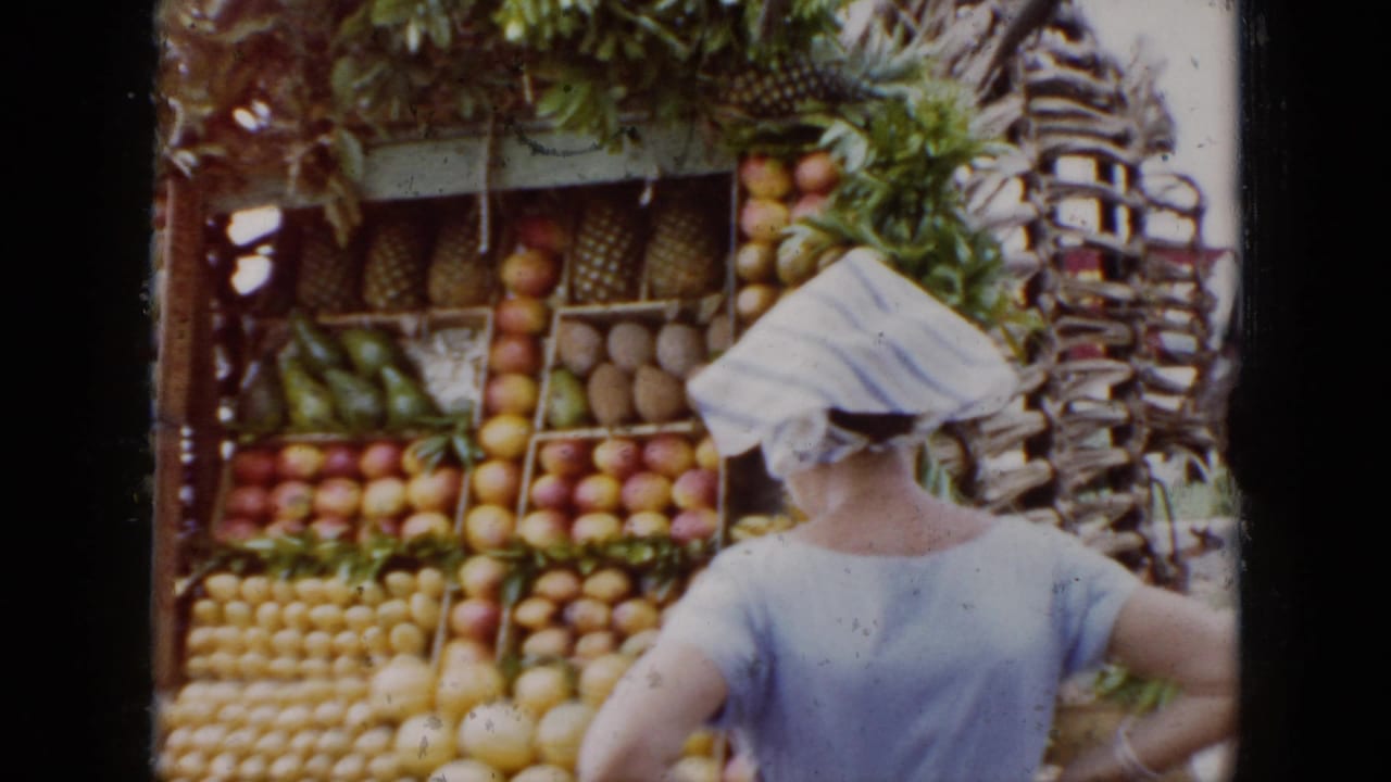 In 1956 Cuba, a woman in a white headscarf appears near stacked tropical fruit at a roadside stand.