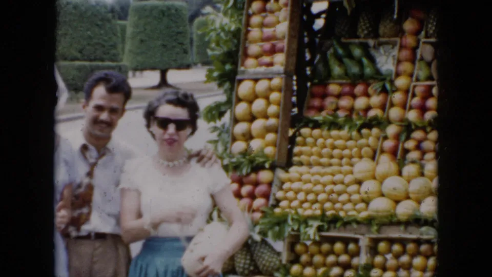 A man in a tie and a woman in sunglasses pose in front of a roadside fruit stand in 1956 Cuba.