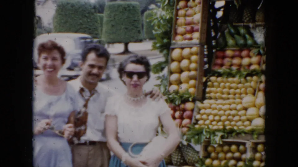 Three adults pose by a Cuba roadside fruit stand, with stacked produce and a classic car in the background.