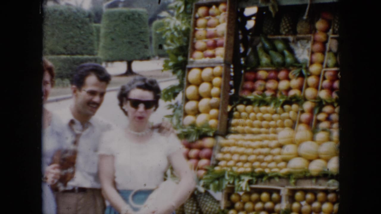The 1956 Cuba home-movie frame shows two adults near stacked oranges, apples, melons, and bananas at a roadside stand.