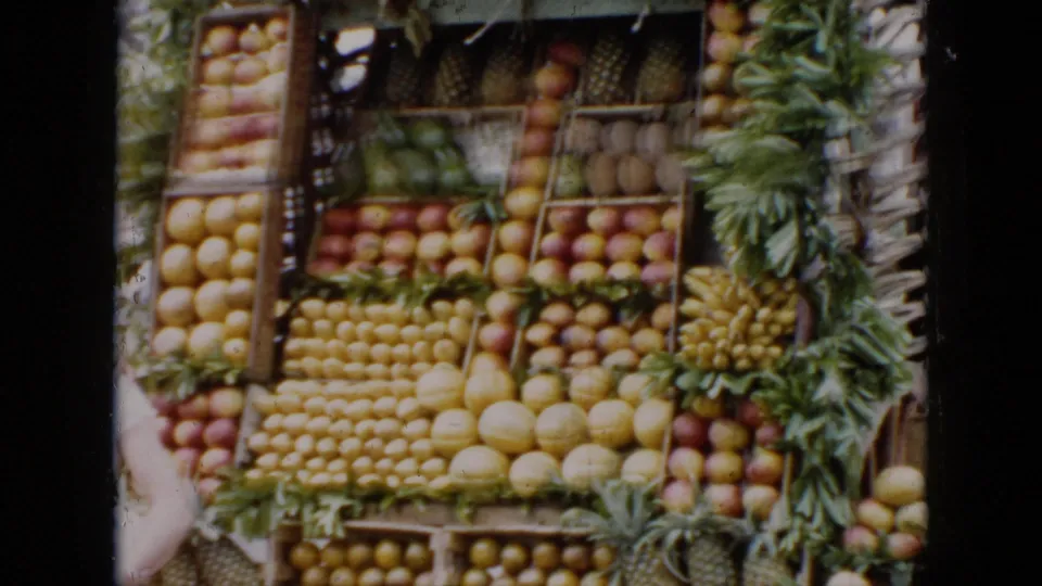 A closer 1956 Cuba frame centers the tiered roadside fruit display, with one person partly visible at the edge.