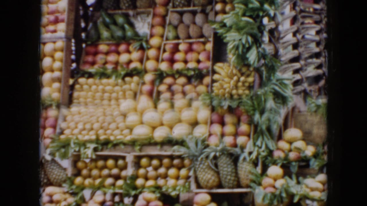 1956 Cuba 8mm frame showing a market fruit display arranged in wooden crates with pineapples, citrus, bananas, and melons.