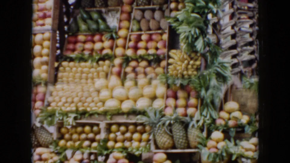 1956 Cuba 8mm frame showing a market fruit display arranged in wooden crates with pineapples, citrus, bananas, and melons.