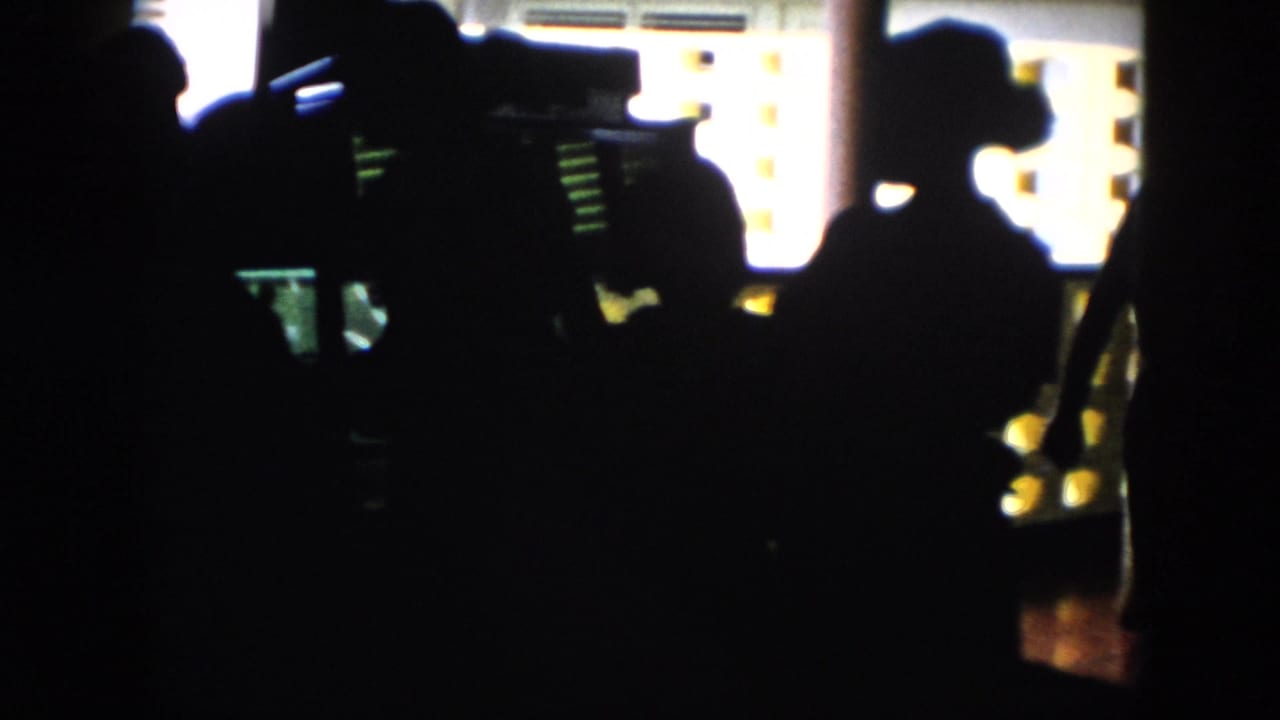 In 1956 Cuba home-movie footage, silhouetted figures gather in a dim interior with bright patterned wall openings behind them.