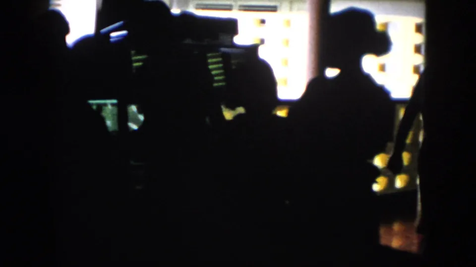 In 1956 Cuba home-movie footage, silhouetted figures gather in a dim interior with bright patterned wall openings behind them.