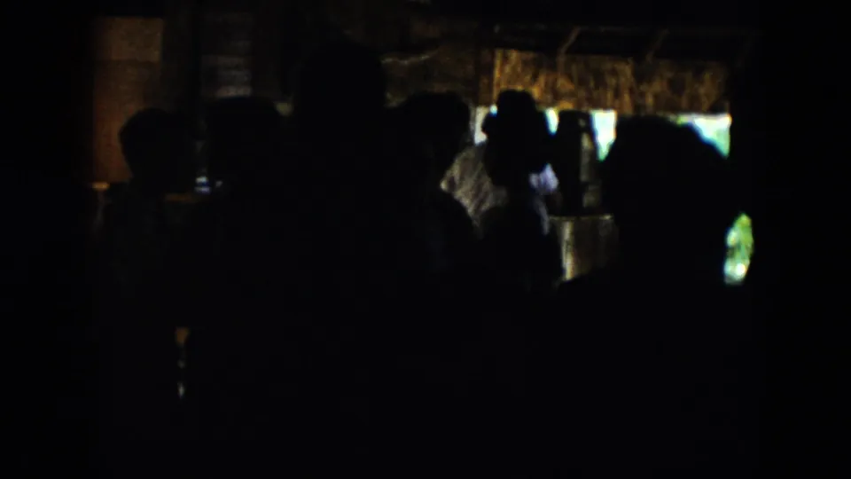 A shadowed group gathers inside, with windows and brighter exterior light visible beyond the silhouettes.