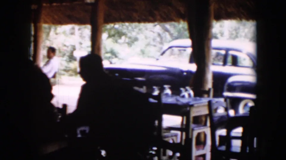 Inside the shaded Cuba 1956 gathering space, chairs and a table sit near the opening while a classic car is parked outside.