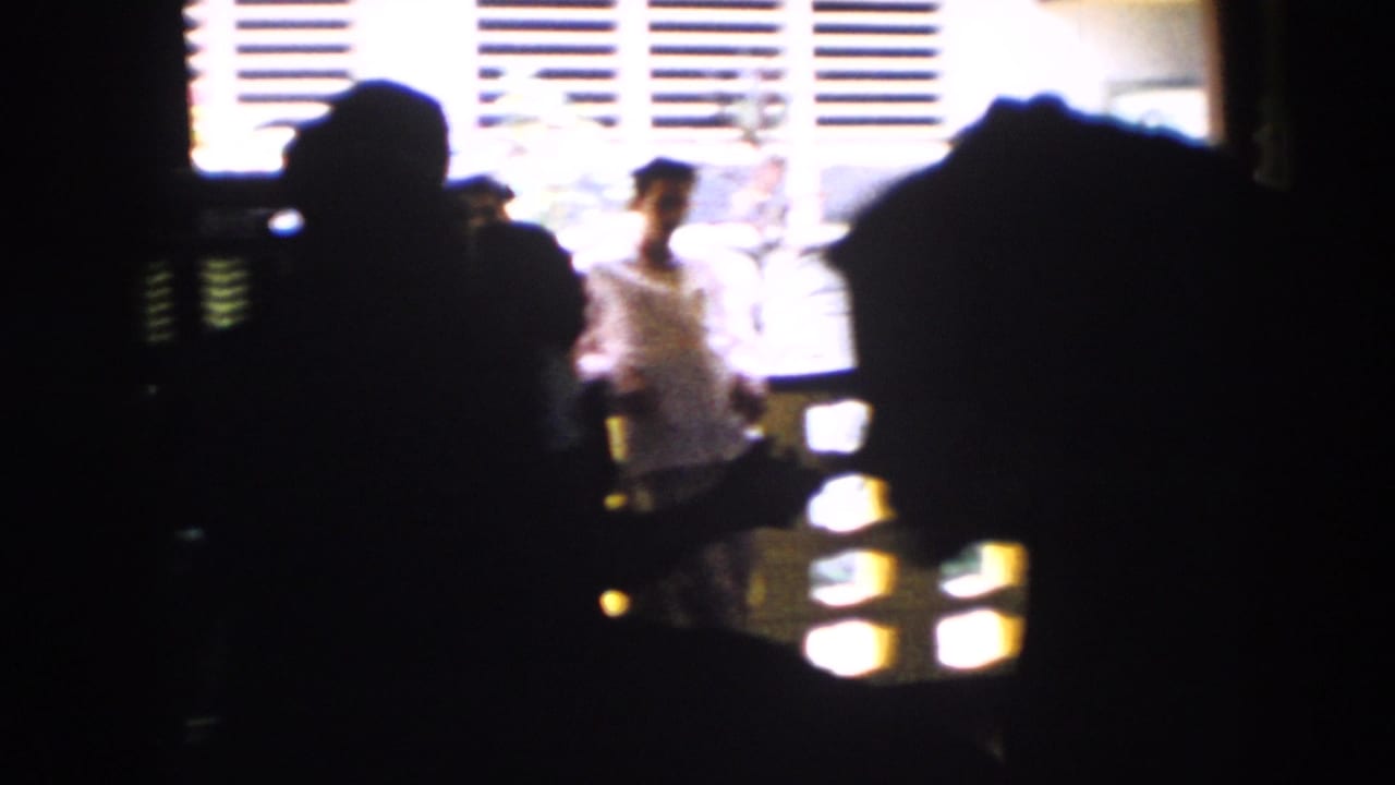 A dim 1956 Cuba home-movie view shows partygoers in silhouette, with one light-clothed person near bright window blinds.