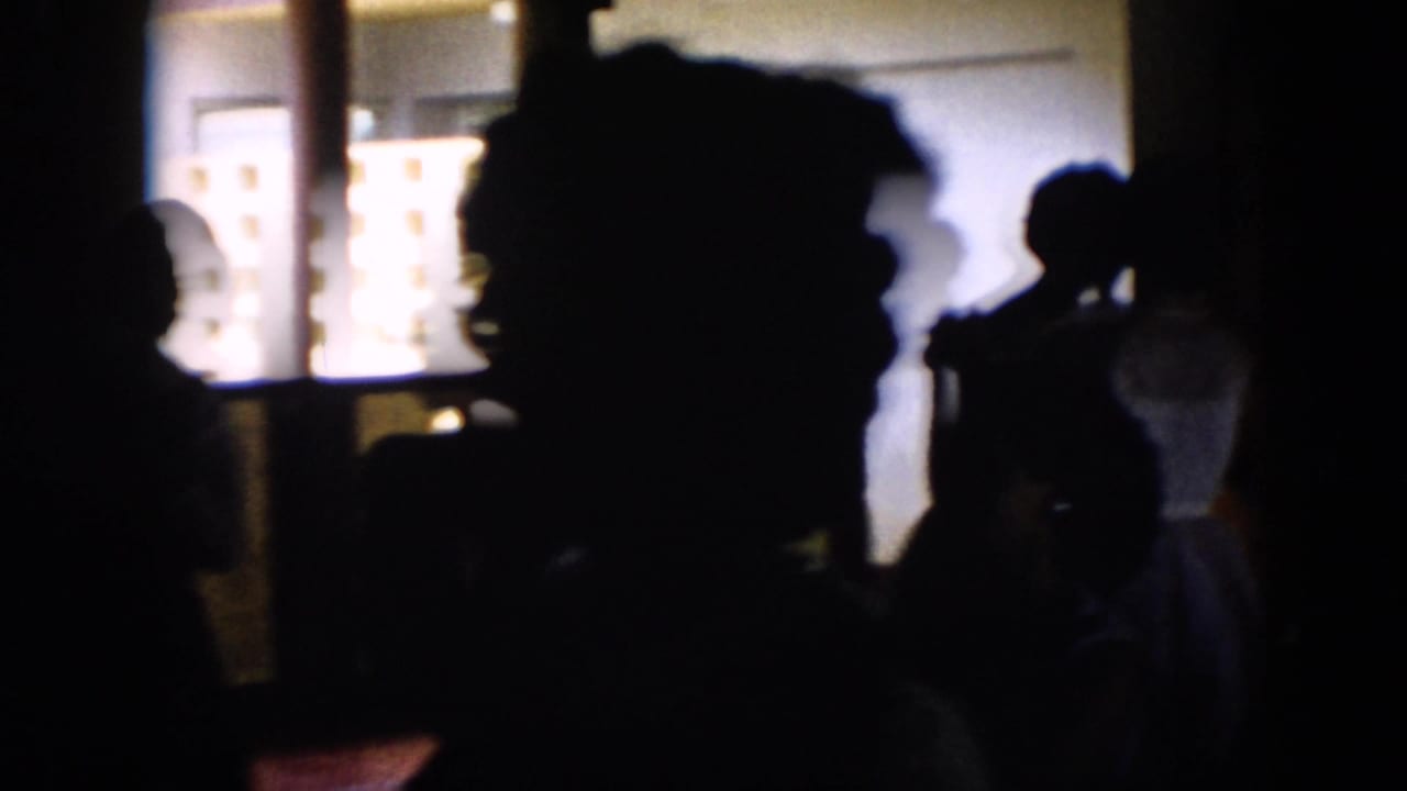 Silhouetted dancers or guests fill the foreground of a dim Cuba party hall, backed by bright windows and interior wall panels.