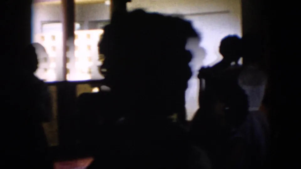 Silhouetted dancers or guests fill the foreground of a dim Cuba party hall, backed by bright windows and interior wall panels.