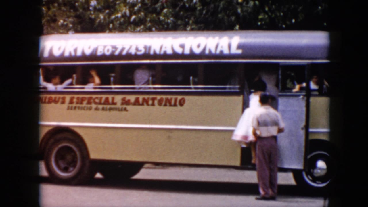 1956 Cuba home-movie frame showing a cream omnibus with visible lettering, passengers at open windows, and a man standing by the entry door.
